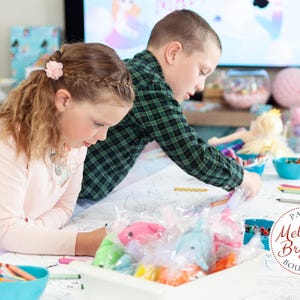 May include: Two children are seated at a table, coloring. The table is covered with a large coloring sheet, crayons, and small bags of colorful items. A logo for "Party Melinda Bryant Boutique" is visible.