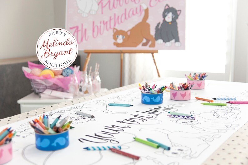 A white table with a beige tablecloth with black polka dots. The tablecloth has a large coloring sheet with a cat theme. There are several small containers of crayons on the table. The text on the coloring sheet says "Your text here". In the background there's a personalized kitten poster -- also available.