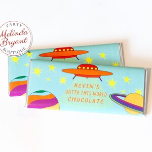 Könnte beinhalten: Zwei blaue und weiße Schokoladenriegelverpackungen mit Weltraumthema. Die Verpackungen zeigen ein rotes und oranges Raumschiff, einen Planeten mit Ringen und Sterne. Der Text auf der Verpackung lautet "Kevin's Outta This World Chocolate".