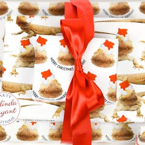 May include: White wrapping paper with a repeating pattern of bearded dragons wearing Santa hats. The text "Merry Christmas Hannah!" is printed on the paper. A red satin ribbon is tied around the paper.