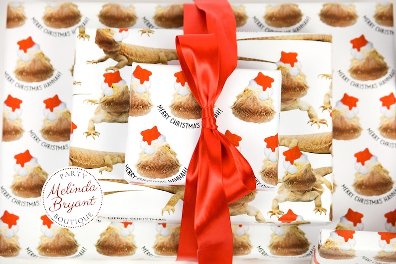 May include: White wrapping paper with a repeating pattern of bearded dragons wearing Santa hats. The text "Merry Christmas Hannah!" is printed on the paper. A red satin ribbon is tied around the paper.
