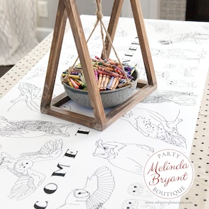 May include: A white table runner with an owl theme, featuring the words "Come" and "Party Melinda Bryant Boutique" printed on it. A wooden stand with a metal bowl filled with crayons hangs above the table runner.