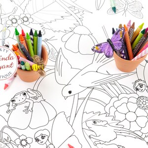 This detail image of my fairy coloring table runner features a bird flying over a garden filled with fairies, flowers, grass, and small creatures including a frog and a chipmunk. It is staged with two small terracotta pots filled with crayons.
