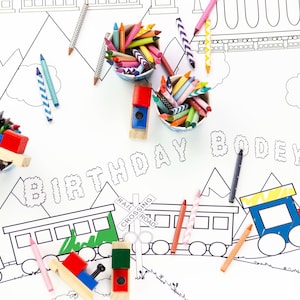 Puede incluir: Un mantel blanco para colorear con temática de tren. El mantel presenta un tren, montañas y las palabras "2nd Birthday Bodey!" El mantel está rodeado de lápices de colores en vasos pequeños.