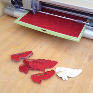 May include: A silver electronic cutting machine with a green mat holding a sheet of red glitter material. Several red glitter shoe-shaped cutouts are stacked on the surface, alongside a few plain white shoe-shaped cutouts.