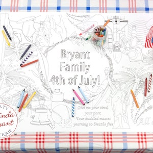 May include: A black and white coloring page with a patriotic theme. The page features a wreath with the text "Bryant Family 4th of July!" inside. The page also includes images of fireworks, the Liberty Bell, a baseball, and a bald eagle. There are also crayons and jars of crayons on the table.