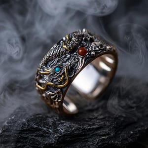 May include: A silver ring with a detailed dragon design. The dragon has gold accents and is decorated with a turquoise and a red gemstone. The ring has an open design and is set against a dark, smoky background.