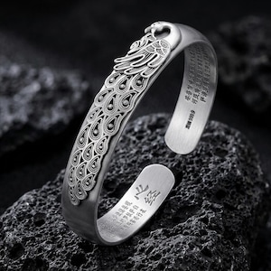 May include: A silver cuff bracelet with a peacock design. The bracelet has an open design with a peacock head and tail feathers. The feathers are detailed with intricate patterns. The bracelet is resting on a dark, textured surface.