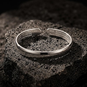Puede incluir: Un brazalete abierto plateado con caracteres grabados. El brazalete se exhibe sobre una superficie oscura y texturizada. El brazalete está marcado con el número "999".