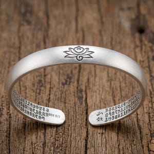 Peut inclure: Bracelet manchette argenté avec un motif de fleur de lotus gravé au centre. Le bracelet présente du texte gravé sur les bords intérieurs. Le bracelet est ouvert en bas.