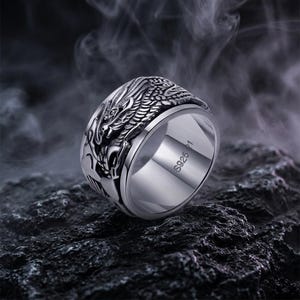 Può includere: Un anello d'argento con un design dettagliato di drago. L'anello ha una fascia rotante e il testo "S925 21" è visibile. L'anello è posto su uno sfondo scuro e strutturato.