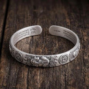Könnte beinhalten: Ein silbernes Armreif-Armband mit aufwendigen geprägten Designs. Das Armband zeigt chinesische Schriftzeichen und Symbole, darunter Münzen und ein Fabelwesen. Das offene Design ermöglicht ein einfaches Tragen. Das Armband ist aus Silber.