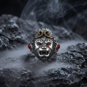 Peut inclure: Bague en argent représentant un visage de singe détaillé, avec des yeux en pierres rouges et des accents dorés. La bague a une finition texturée. La bouche du singe est ouverte, révélant les dents. Sur fond sombre et rocheux.