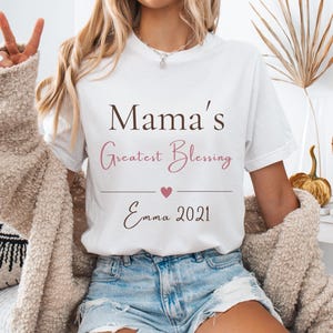 Könnte beinhalten: Ein weißes Kurzarm-T-Shirt mit dem braunen Schriftzug Mama's, dem rosafarbenen Schreibschrift-Text Greatest Blessing, einem kleinen rosa Herz und dem Text Emma 2021 in Braun. Das T-Shirt hat einen Rundhalsausschnitt und eine normale Passform.