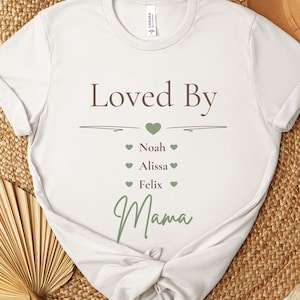 Könnte beinhalten: Ein weißes Kurzarm-T-Shirt mit dem braunen Schriftzug "Loved By", gefolgt von einem grünen Herz, den Namen "Noah", "Alissa" und "Felix", die jeweils von kleinen grünen Herzen umgeben sind, und dem Wort "Mama" in grüner Schreibschrift.
