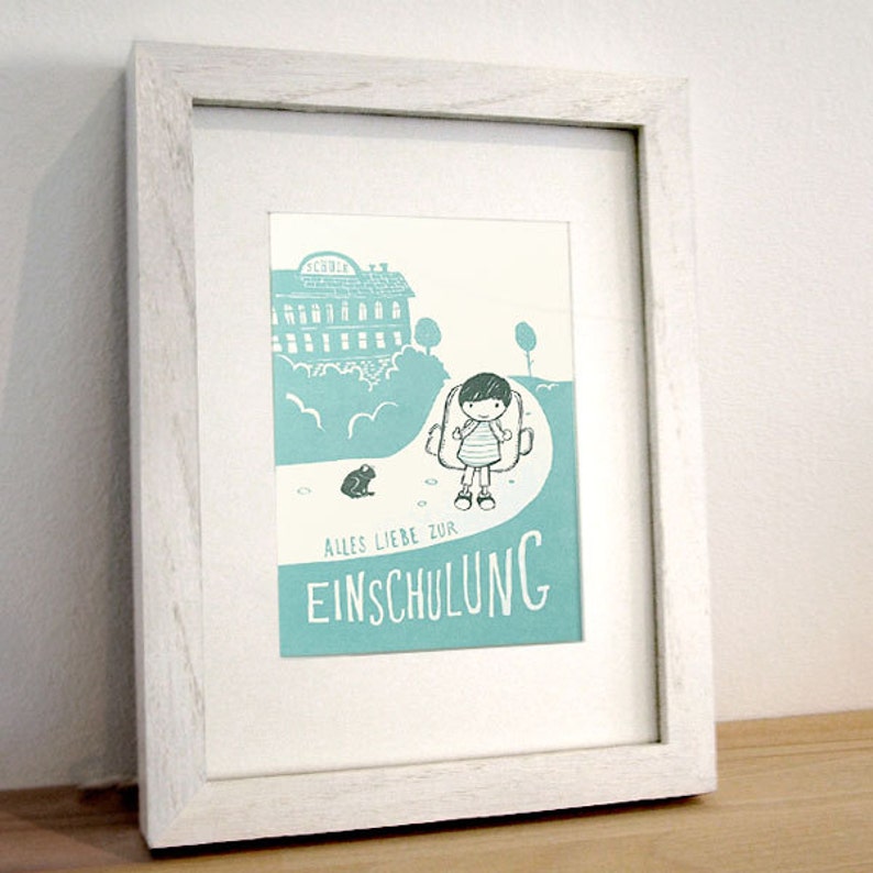 May include: A white framed print with a light blue background. The print features a cartoon illustration of a child with a backpack walking towards a school building. The text "Alles Liebe zur Einschulung" is written in a decorative font.