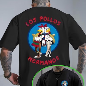 Peut inclure: T-shirt noir avec une image de dessin animé de deux poulets portant des sombreros, avec le texte "LOS POLLOS HERMANOS" en rouge. L'image est dans un cercle bleu. Col rond.