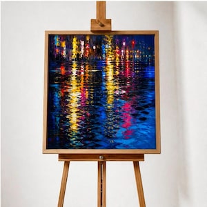 May include: A framed painting on a wooden easel depicts a nighttime cityscape reflected in water. The artwork features vibrant colors, including yellow, red, and blue, creating a dynamic and colorful scene. The wooden frame and easel add a natural touch.