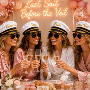 Może przedstawiać: Cztery białe czapki kapitańskie z czarnymi paskami i złotym haftem, każda z unikalnym napisem, w tym Captain Emma, Crew, Bride i Captain Olivia. Czapki noszone są do różowych satynowych szlafroków i ciemnych okularów przeciwsłonecznych.