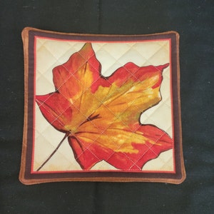 May include: A quilted brown and cream hot pad with a large red and yellow maple leaf design.