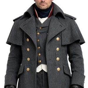 Könnte beinhalten: Ein grauer Wollmantel mit goldenen Knöpfen, eine passende Weste und schwarze Lederhandschuhe. Der Mantel hat einen Schulterkragen und wird mit schwarzen Hosen und Stiefeln getragen. Ein schwarzer Zweispitz rundet das historische, militärisch inspirierte Ensemble ab.