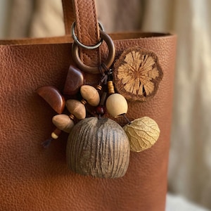 May include: A brown leather tote bag with a decorative charm. The charm features a variety of natural elements, including wooden beads, a textured seed pod, and a dried flower. The bag has a simple design with a handle and a metal ring.