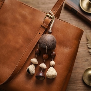May include: A brown leather tote bag with a decorative charm. The charm is made of wood and beads in shades of brown and beige. The bag has a shoulder strap and a metal buckle.
