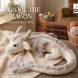May include: A crocheted dragon toy in white and gray, lying on a cream-colored blanket. The dragon has horns, wings, and a long tail. The text "WINNIE THE DRAGON CROCHET PATTERN" is at the top. The pattern difficulty is intermediate.