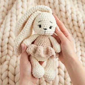May include: A handmade crochet bunny doll in cream and tan. The bunny has long, floppy ears, a small face with black embroidered eyes and a stitched nose. It wears a tan dress. The doll is held in two hands against a cream background.