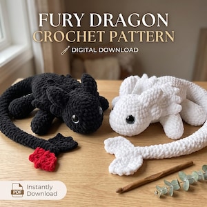 May include: A digital download crochet pattern titled FURY DRAGON CROCHET PATTERN featuring two plush dragon toys. One dragon is black with a red tail tip, and the other is white. Both have large dark eyes and textured yarn bodies.