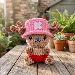May include: A crocheted plush toy featuring a tan creature wearing a pink bucket hat with a white cross emblem. The figure has brown antlers, black eyes, a blue nose, pink cheeks, and is wearing red shorts. The texture is soft and fuzzy.