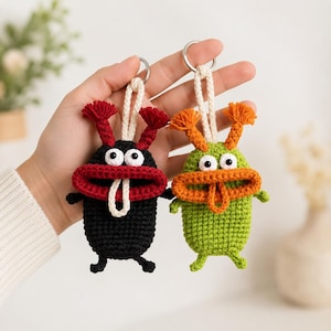 Op de afbeelding: Twee gehaakte monster-sleutelhangers met metalen ringen. De ene is zwart met een donkerrode mond en antennes, de andere is limoengroen met een oranje mond en antennes. Beide hebben witte ogen en een bungelende tong van wit garen.