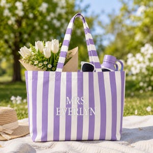 May include: A purple and white striped canvas tote bag featuring the embroidered text MRS. EVERLYN in white. The bag contains a bouquet of white tulips and daisies in a brown paper wrap, a pair of black sunglasses, and a purple water bottle.