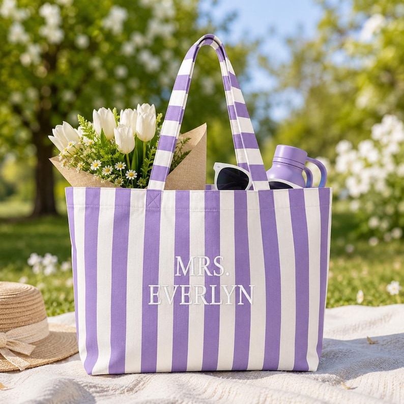 May include: A purple and white striped canvas tote bag featuring the embroidered text MRS. EVERLYN in white. The bag contains a bouquet of white tulips and daisies in a brown paper wrap, a pair of black sunglasses, and a purple water bottle.