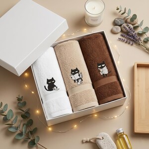 K&ouml;nnte beinhalten: Eine wei&szlig;e Geschenkbox enth&auml;lt drei gerollte Handt&uuml;cher in Wei&szlig;, Beige und Braun. Jedes Handtuch hat ein schwarzes Katzenmotiv. Das wei&szlig;e Handtuch zeigt eine sitzende Katze, das beige Handtuch eine lesende Katze und das braune Handtuch eine in ein Handtuch gewickelte Katze.