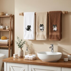 K&ouml;nnte beinhalten: Drei Badet&uuml;cher mit schwarzer Katzenstickerei, die an einer Holzstange h&auml;ngen. Die Handt&uuml;cher sind wei&szlig;, beige und braun. Das wei&szlig;e Handtuch zeigt eine lesende Katze, das beige Handtuch eine Katze, die ein Buch liest, und das braune Handtuch eine Katze, die in ein Handtuch gewickelt ist.