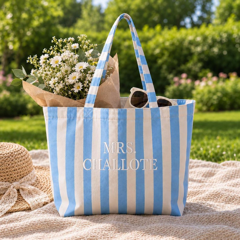 May include: A tote bag featuring vertical light blue and white stripes with the embroidered text MRS. CHALLOTE in white. The bag has two matching striped shoulder straps and contains a bouquet of white flowers and a pair of dark sunglasses.