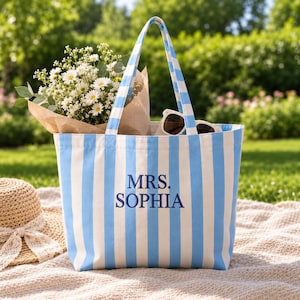 May include: A light blue and white vertical striped canvas tote bag featuring the embroidered text MRS. SOPHIA in dark blue thread. The bag has two matching striped shoulder straps and is filled with a bouquet of white flowers and sunglasses.