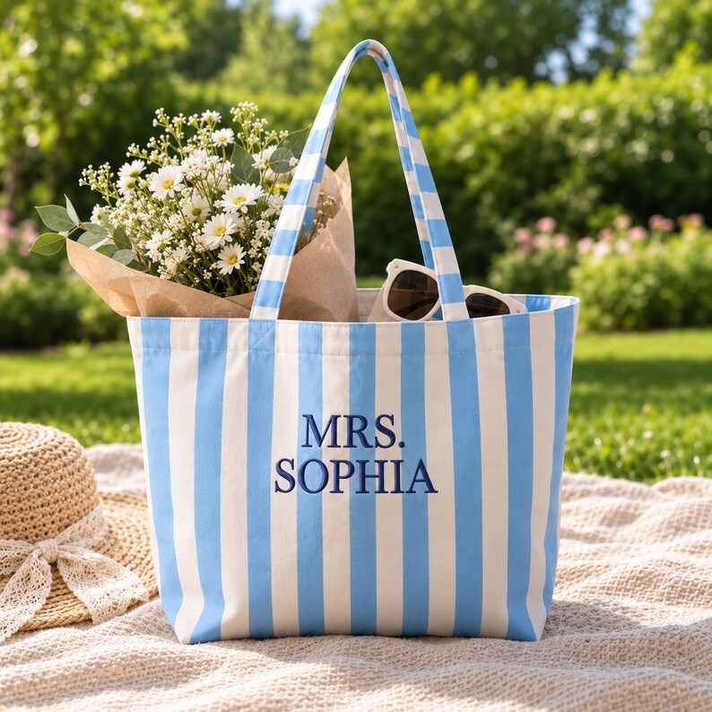 May include: A light blue and white vertical striped canvas tote bag featuring the embroidered text MRS. SOPHIA in dark blue thread. The bag has two matching striped shoulder straps and is filled with a bouquet of white flowers and sunglasses.