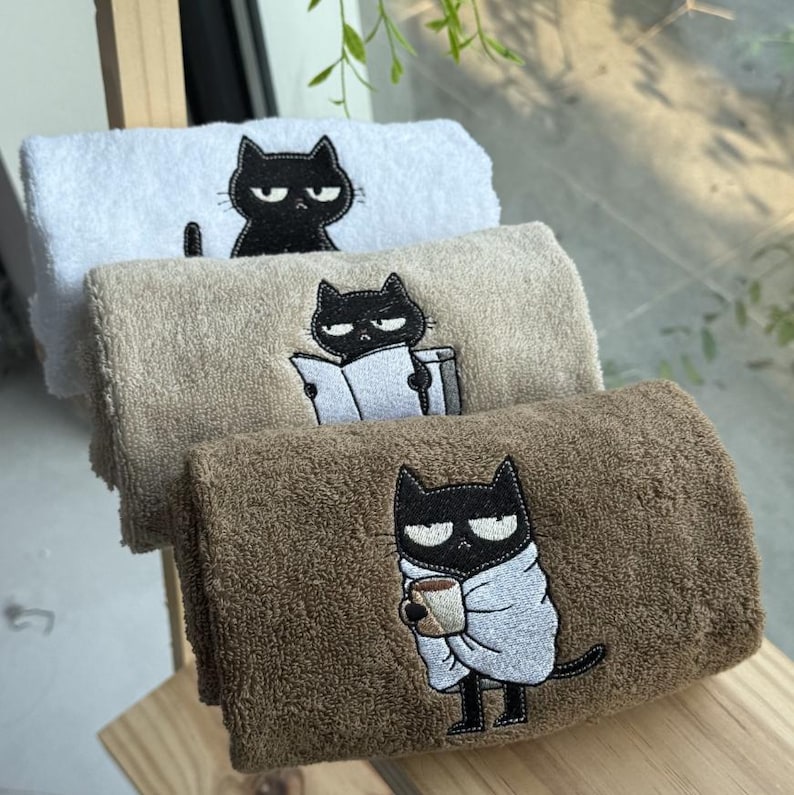 K&ouml;nnte beinhalten: Drei gerollte Handt&uuml;cher mit gestickten schwarzen Katzenmotiven. Das obere Handtuch ist wei&szlig;, das mittlere ist beige und das untere ist braun. Jede Katze hat eine andere Pose, darunter eine, die liest und eine andere, die eine Tasse h&auml;lt.