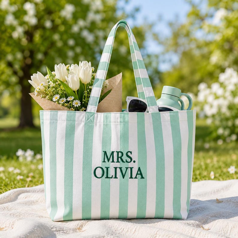 May include: A tote bag featuring vertical mint green and white stripes with the text MRS. OLIVIA embroidered in dark green. The bag contains a bouquet of white tulips wrapped in brown paper and a light green water bottle with a handle.