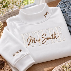 Puede incluir: Una sudadera blanca con bordados en marrón y blanco. El cuello muestra J corazón T, el pecho dice FUTURE Mrs. Smith en letras cursivas y de molde superpuestas, y el puño de la manga tiene bordada la fecha 12.06.26.