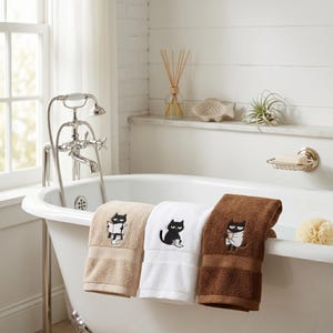 K&ouml;nnte beinhalten: Drei Badet&uuml;cher in Beige, Wei&szlig; und Braun, jedes mit einem schwarzen Katzenmotiv bestickt. Die Handt&uuml;cher sind &uuml;ber den Rand einer wei&szlig;en Badewanne gefaltet. Das Badezimmer hat ein Fenster, einen Wasserhahn und ein Regal mit dekorativen Gegenst&auml;nden.