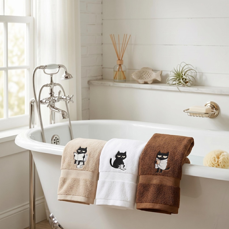 K&ouml;nnte beinhalten: Drei Badet&uuml;cher in Beige, Wei&szlig; und Braun, jedes mit einem schwarzen Katzenmotiv bestickt. Die Handt&uuml;cher sind &uuml;ber den Rand einer wei&szlig;en Badewanne gefaltet. Das Badezimmer hat ein Fenster, einen Wasserhahn und ein Regal mit dekorativen Gegenst&auml;nden.