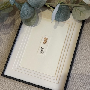 Puede incluir: Una obra de arte enmarcada que presenta dos pequeñas ilustraciones rectangulares estilo sello postal centradas en un paspartú color crema. El sello superior representa un perro marrón, mientras que el inferior muestra un perro blanco y negro.
