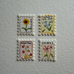 Op de afbeelding: Vier rechthoekige illustraties in postzegelstijl met handgeschilderde botanische ontwerpen. Linksboven een gele zonnebloem met het label SUNFLOWER. Rechtsboven blauwe bloemen met het label FORGET ME NOT. Linksonder rode tomaten met het label TOMATOS. Rechtsonder roze cosmosbloemen met het label COSMOS.