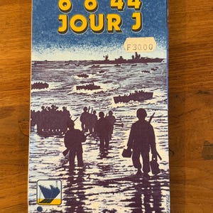 Può includere: Scatola di gioco da tavolo vintage con sfondo blu e testo "D DAY 6 6 44 JOUR J" in giallo. La scatola presenta una grafica di soldati che guadano nell'acqua verso una spiaggia, con navi sullo sfondo. Il logo di IGN France è in basso.