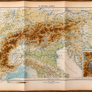 Può includere: Una mappa fisica vintage intitolata IL SISTEMA ALPINO, che mostra un rilievo topografico dettagliato delle Alpi in tonalità marroni, verdi e blu. La mappa include un riquadro più piccolo della regione del Monte Bianco e una scala grafica.