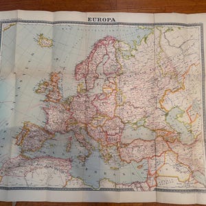 Può includere: Una mappa d'epoca dell'Europa, intitolata "Europa", con dettagli geografici e confini dei paesi. La mappa è stampata su carta invecchiata, con una tavolozza di colori beige, rosso, verde e blu. La mappa è piegata e appoggiata su una superficie di legno.