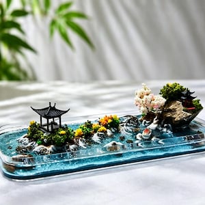 May include: A miniature Asian-inspired diorama featuring a black pagoda, colorful flowers, and a small boat set on a blue water base. The scene includes a small island with a red-roofed structure and a rock formation.